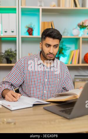 L'uomo indiano sul tavolo di casa legge i quaderni e scrive appunti mentre studia durante le lezioni online a casa. L'arabo sul tavolo di casa controlla fatti e studi con particolare attenzione all'apprendimento online remoto. Foto Stock
