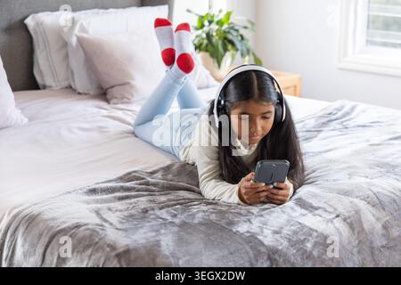 Ragazza indiana in età scolare sdraiata sul letto di casa con cuffie, smartphone, calzini rossi Foto Stock