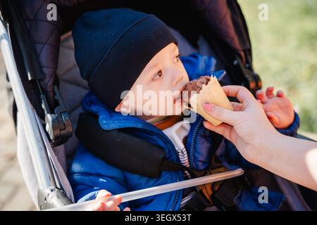 Bambino che mangia gelato al cioccolato in un passeggino indossando una giacca blu e un cappello nero all'aperto alla luce del sole. Stile di vita infantile. Foto Stock