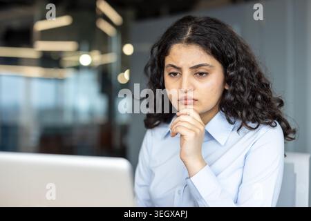 Giovane donna con capelli ricci scuri che indossa una camicia azzurra che si concentra sullo schermo di un notebook, che pensa e risolve un problema in un ufficio moderno Foto Stock