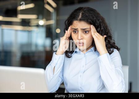 Giovane donna indiana che stringe la testa in un ufficio moderno, stressata e affaticata mentre lavora su un notebook, raffigurante ustione, ansia e pressione sul posto di lavoro Foto Stock