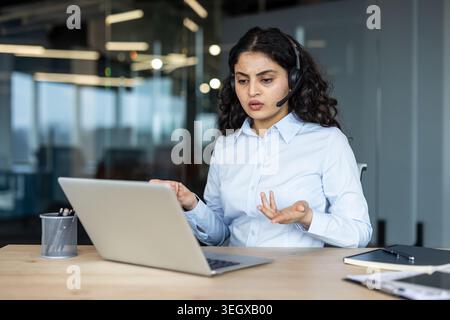 Una donna in camicia aziendale che indossa un auricolare partecipa a una riunione virtuale su un laptop, gestendo attivamente mentre fornisce assistenza o assistenza clienti in un ambiente di ufficio moderno Foto Stock