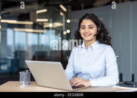 Giovane donna sorridente che indossa un auricolare e digita un laptop in un ufficio moderno, fornendo assistenza clienti e consulenza online in qualità di operatore remoto e consulente professionista Foto Stock