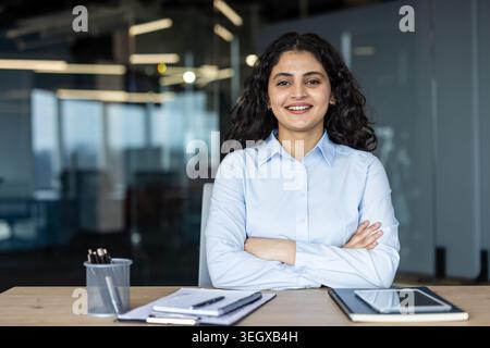 Giovane donna indiana professionale con capelli ricci seduta alla scrivania, sorridendo con fiducia e guardando la telecamera, simboleggiando il successo di carriera e la leadership aziendale Foto Stock