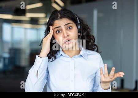 Giovane agente dell'assistenza clienti che mostra shock e frustrazione durante una chiamata difficile, mano sulla testa, bocca aperta, indossa l'auricolare e camicia azzurra in un ritratto dell'ufficio Foto Stock