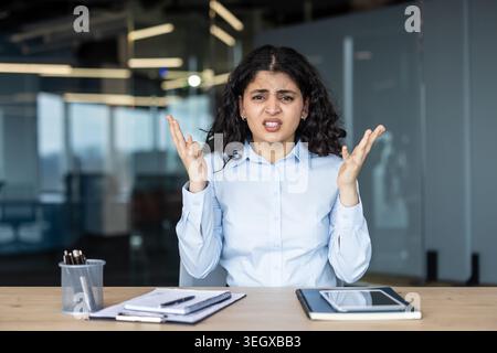 Una giovane lavoratrice d'ufficio siede alla scrivania con le mani alzate, esprimendo frustrazione, confusione e stress durante una giornata lavorativa tesa in un moderno ambiente di lavoro aziendale, ritratto Foto Stock