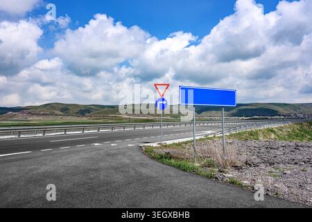 Il cartello stradale si trova accanto alla strada asfaltata vuota. Segni rettangolari blu e triangolo rosso visibili. Cielo con soffici nuvole bianche Foto Stock