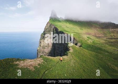 Veduta aerea di un escursionista con una giacca arancione che cammina lungo le spettacolari scogliere verdi della riserva naturale di Hornstrandir, affacciato sull'oceano blu di Isafjordu Foto Stock