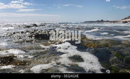 Le onde baciate dal sole dell'oceano Atlantico creano delicatamente schiuma bianca all'interno di una piscina rocciosa con marea al largo di una spiaggia sabbiosa sotto un cielo blu e nuvoloso. Foto Stock