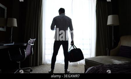 Giovane uomo d'affari con borsa da viaggio in piedi vicino a una grande finestra in una moderna camera d'albergo mentre controlla lo smartphone. Manager che monitora le informazioni sui dispositivi mobili Foto Stock