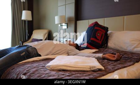 Uomo esausto sdraiato sul letto in una camera d'albergo moderna dopo un lungo viaggio d'affari. Giovane uomo d'affari stanco che si rilassa in una sistemazione confortevole dopo Foto Stock