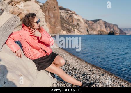 Giacca rosa donna Coast giovane donna in una giacca rosa si rilassa su una spiaggia rocciosa vicino al mare blu Foto Stock