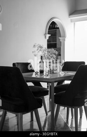 Ampia immagine monocromatica degli interni contemporanei della sala da pranzo, che mostra sedie rivestite scure intorno a un tavolo fisso con vino, bicchieri e fiori, cornice Foto Stock