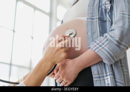 Medico che esamina la donna incinta che tiene lo stetoscopio vicino alla pancia grande che ascolta il battito cardiaco del bambino. Ginecologo medico consulenza paziente su gravidanza in ufficio ospedaliero. Visita al controllo medico medico Foto Stock