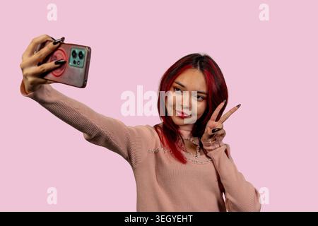 Una giovane donna con i capelli rossi mette in risalto un autoritratto sul telefono su uno sfondo rosa Foto Stock