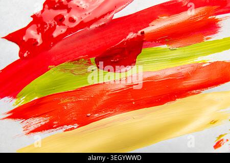 Le strisce di vernice rossa, verde e gialla si mescolano su una tela bianca nello studio di un artista sotto la luce naturale durante il giorno. Foto Stock