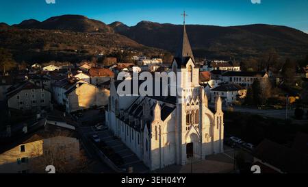 Vista aerea dell'Eglise Saint-Pierre con la sua alta guglia e la facciata in pietra circondata dai tetti e dalle montagne del Giura al tramonto a Gex, Auvergne-R. Foto Stock