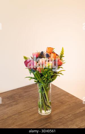 Un bouquet pieno di diversi tipi di fiori sorge in un vaso di vetro trasparente su un tavolo di legno. L'impostazione è in interni con uno sfondo chiaro. Foto Stock