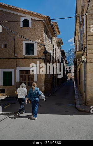 Maiorca, Spagna - 21 maggio 2026: Due donne si allontanano lungo uno stretto vicolo illuminato dal sole in una storica città di pietra, incorniciata da finestre con persiane, lampade e m Foto Stock