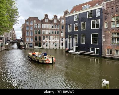 Vista di una barca con persone che navigano lungo i canali di Amsterdam circondata da case strette storiche con facciate a spiovente nei Paesi Bassi. Foto Stock