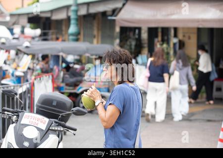 BANGKOK, THAILANDIA - 16 MAGGIO 2026: Turista femminile che beve acqua fresca di cocco, vita quotidiana nel mercato asiatico di strada, profilo laterale Foto Stock