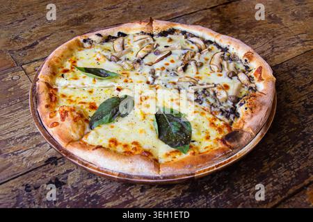 La pizza appena sfornata su un tavolo di legno è abbondantemente condita con mozzarella fusa funghi saltati e foglie di basilico fresco, serviti su una b Foto Stock