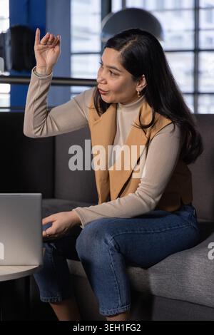 Donna asiatica che indossa un gilet beige sopra il dolcevita, seduta sul divano in ufficio mentre gesti vicino al computer portatile Foto Stock