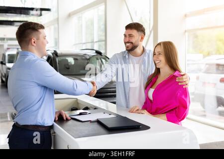 Coppia felice che stringe la mano al venditore di auto presso la concessionaria dopo l'acquisto del veicolo Foto Stock