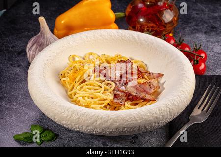 Deliziosi spaghetti alla carbonara con pancetta croccante e formaggio in una ciotola rustica bianca. Ingredienti freschi come aglio, peperone e pomodori circondano il dis Foto Stock