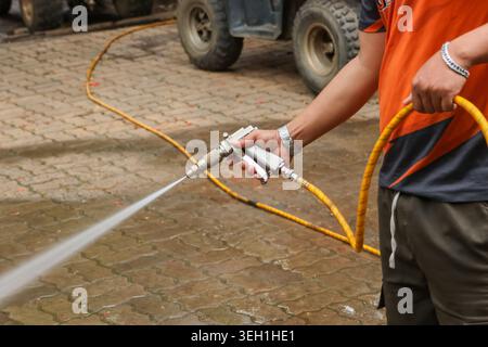 Il lavoratore concentrato gestisce la pulizia della pavimentazione con getto d'acqua dal tubo flessibile giallo durante la manutenzione all'aperto. Man lavaggio superficie vialetto d Foto Stock