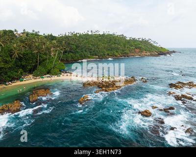 Vista aerea di Secret Beach Mirissa con onde turchesi che si infrangono contro gli affioramenti rocciosi e persone che si rilassano sulla spiaggia sabbiosa di Mirissa, South P. Foto Stock