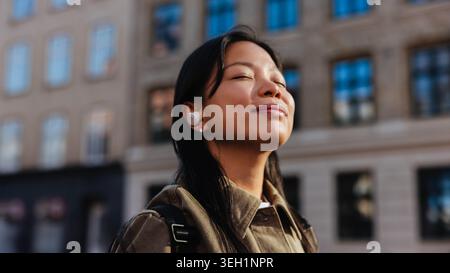 Donna in piedi su una strada della città con gli occhi chiusi e un leggero sorriso, indossa auricolari wireless e uno zaino mentre assorbe la calda luce del sole e ascolta Foto Stock