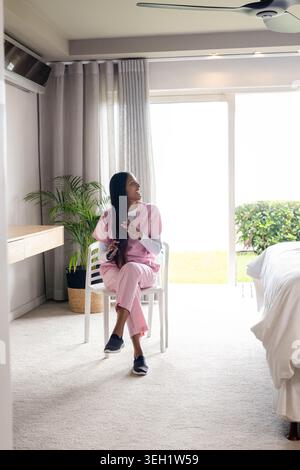 Donna indiana seduta su una sedia bianca in una luminosa camera da letto con scrub rosa che controlla lo smartphone Foto Stock