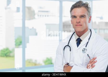 Medico di sesso maschile anziano in piedi presso la finestra della clinica con camice da laboratorio, stetoscopio e penna, spazio di copia Foto Stock