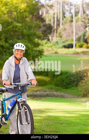 Donna anziana in piedi con una bici blu sorridente nel parco con casco, bottiglia d'acqua, spazio fotocopie Foto Stock