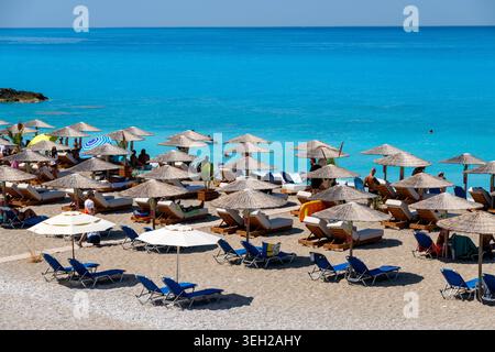 Nydri - Isole Ionie - Grecia - 08-09-2025: File di ombrelloni in paglia e tessuto con lettini da mare, spiaggia sabbiosa accanto al mare turchese, dove le persone si rilassano e nuotano sotto il cielo limpido. Foto Stock