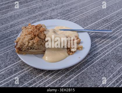 Fetta di torta sbriciolata o crumble di mele con crema pasticcera alla vaniglia su un piatto bianco con cucchiaio, su tovaglia testurizzata grigia, luce naturale all'aperto Foto Stock
