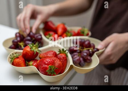Fragole e uva fresche nel vassoio da portata. Primo piano di mani che tengono in mano un vassoio diviso pieno di fragole fresche e uva rossa per una buona salute Foto Stock