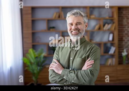 Uomo maturo e sicuro con una camicia casual verde oliva sorridente e guardando la fotocamera, in piedi con le braccia incrociate in un ufficio domestico o in uno spazio abitativo moderno Foto Stock