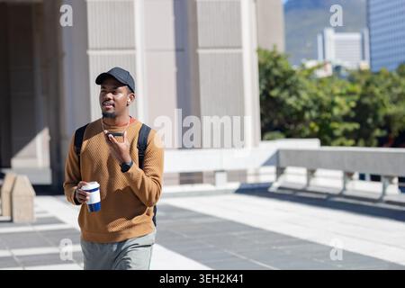 Uomo afroamericano che indossa un berretto che cammina attraverso la piazza tenendo la tazza da viaggio e parlando al telefono Foto Stock