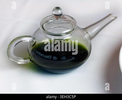 Primo piano di una teiera in vetro trasparente in miniatura ripiena di basilico verde scuro o olio infuso alle erbe, posta su una tovaglia di lino bianco in un raffinato locale da pranzo Foto Stock