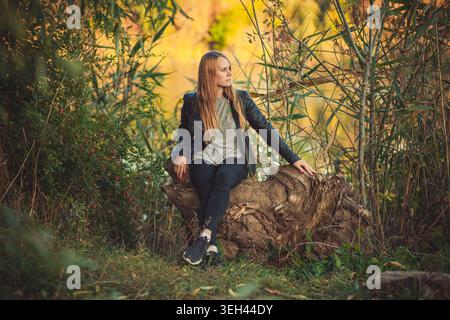 Giovane donna pensiva che si rilassa su un ceppo d'albero nella splendida natura autunnale. Ritratto a figura intera della persona in stile informale, godendosi momenti tranquilli all'aperto. Foto Stock