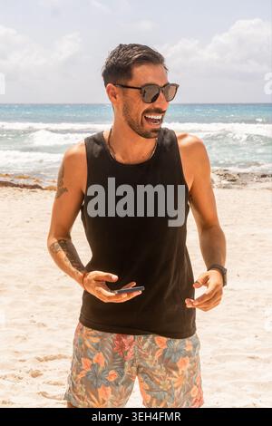 Uomo sorridente tatuato in pantaloncini da surf floreali e canotta nera in piedi su una spiaggia soleggiata, tenendo in mano un telefono e indossando occhiali da sole scuri. Foto Stock