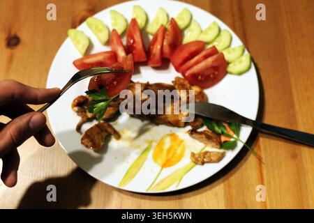 La persona sta tenendo una forchetta con un pezzo di pomodoro e prezzemolo, preparandosi a mangiare una sana cena con pollo arrosto, pomodori a fette e cetriolo Foto Stock