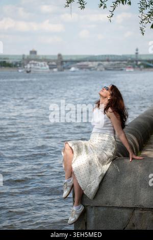 Una donna dai capelli rossi si rilassa sull'argine nel Gorky Park, sul fiume Mosca, sotto la calda luce del sole estivo, sul lungomare urbano. Stile di vita, branding benessere Foto Stock