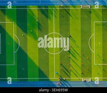 La vista aerea cattura i giocatori che si allenano su un campo da calcio artificiale in erba verde luminoso sotto il sole del tardo pomeriggio che proietta lunghe ombre Foto Stock