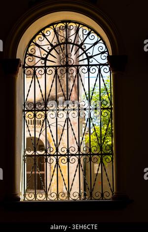 Griglia ornata in ferro battuto che incornicia una luminosa vista del cortile attraverso una finestra ad arco, con forte contrasto tra ombre interne scure e caldo sole Foto Stock