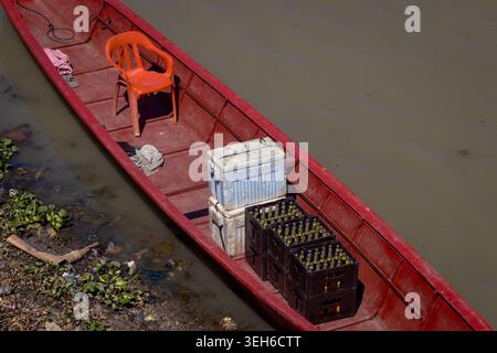 Piccola barca rossa che trasporta casse, refrigeratori e una sedia arancione che galleggia sull'acqua fangosa del fiume, vista dall'alto in un ambiente rustico di trasporto e pesca. Foto Stock