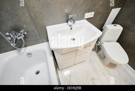 Primo piano di moderni interni del bagno con lavandino, WC e bordo vasca. Bagno pulito con pannelli murali con struttura scura, pavimento in marmo bianco e finiture cromate Foto Stock