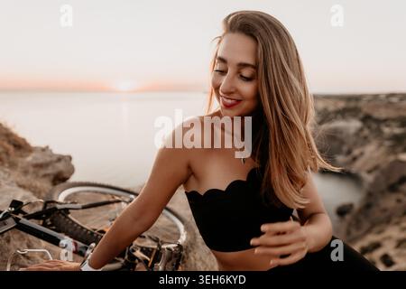 Donna al tramonto, bella giovane donna sorridente mentre si siede con la sua bicicletta sul bordo di una scogliera al tramonto affacciato sul mare Foto Stock